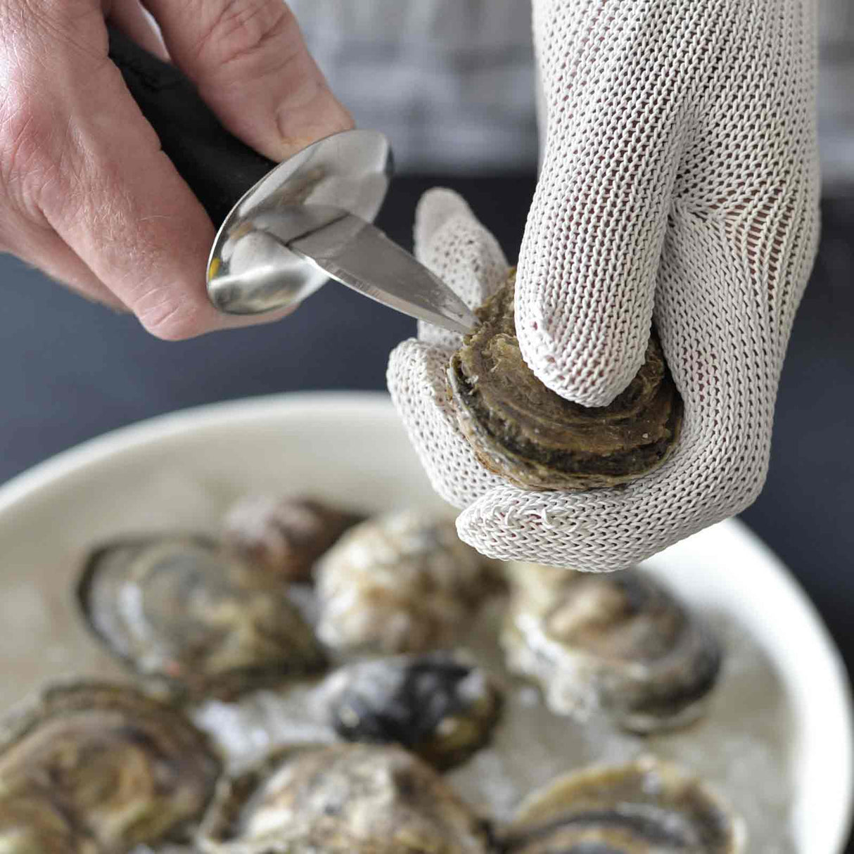 A person wearing a TableCraft cut resistant glove uses an oyster knife to shuck an oyster over a bowl filled with several oysters on ice. Focus is on the hands, glove, knife, and oyster, highlighting the careful shell-opening process.