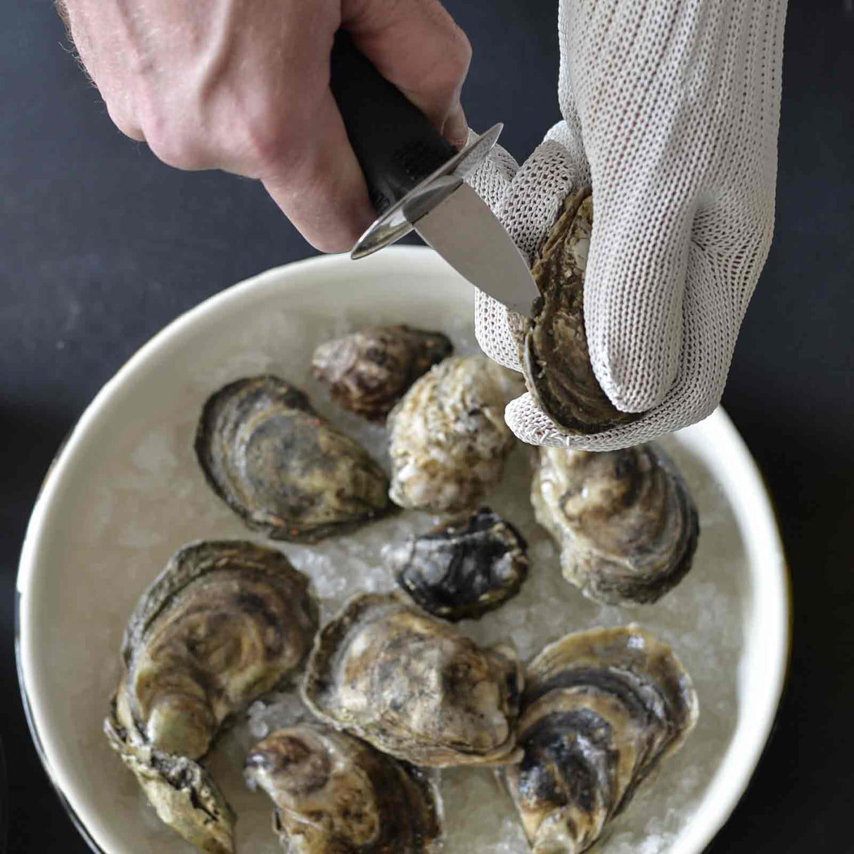 A person uses a knife to shuck an oyster, wearing a cut resistant glove over one hand while holding it above a white plate filled with oysters on ice, set against a dark background.