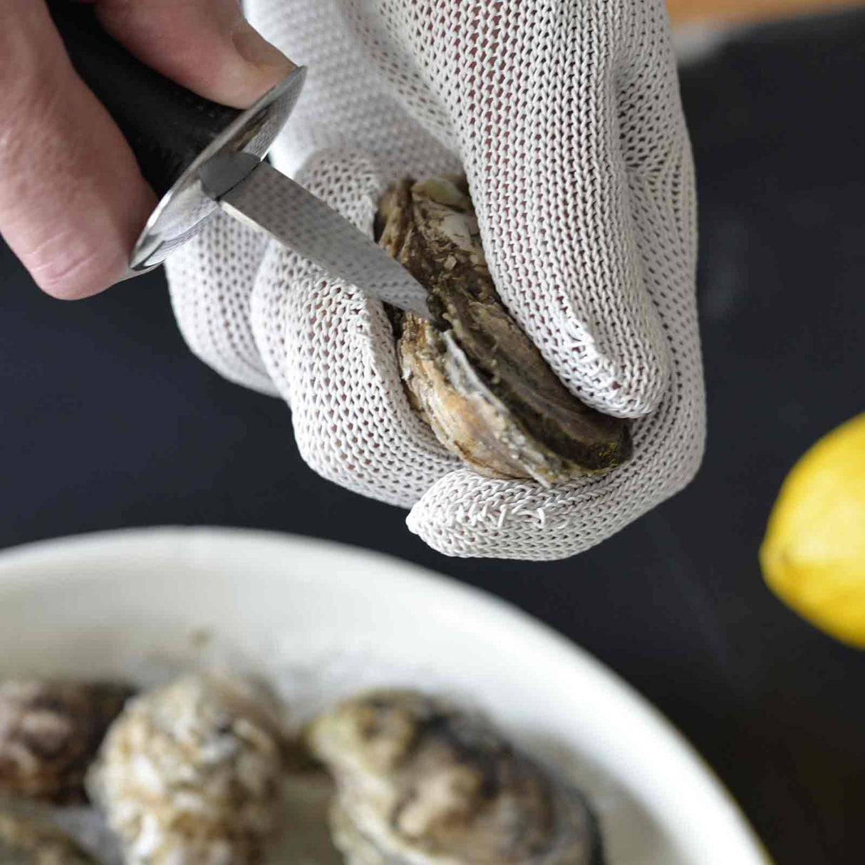 A hand in a cut resistant glove uses an oyster knife to pry open an oyster shell. In the background, a plate with more oysters and a yellow lemon are partially visible. The scene is set on a dark surface.