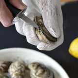 A hand in a cut resistant glove uses an oyster knife to pry open an oyster shell. In the background, a plate with more oysters and a yellow lemon are partially visible. The scene is set on a dark surface.