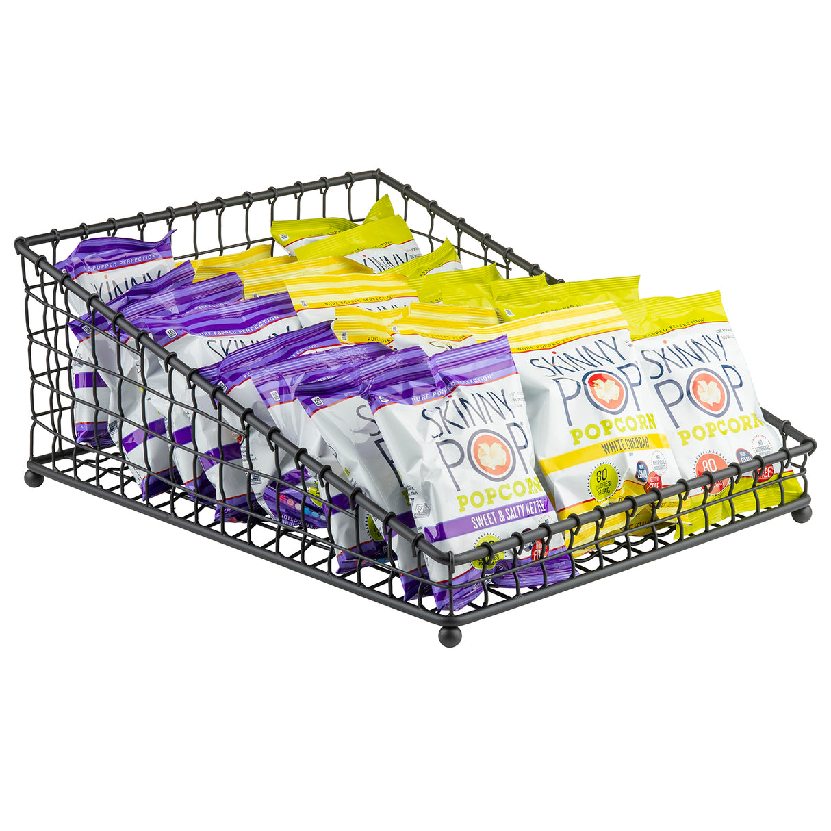 A black powder coated metal Grand Master Collection Basket by TableCraft Products holds several small bags of SkinnyPop popcorn, organized in two rows—white bags with purple accents (Sweet & Salty Kettle) in front and yellow-accented bags in back.