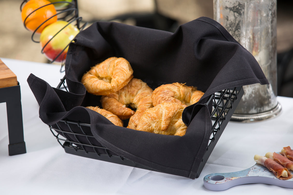 A black powder coated Transformer Basket lined with black cloth holds golden, flaky croissants. The basket sits on a white tablecloth, with a spiral fruit holder in the background and a small charcuterie board partly visible nearby.