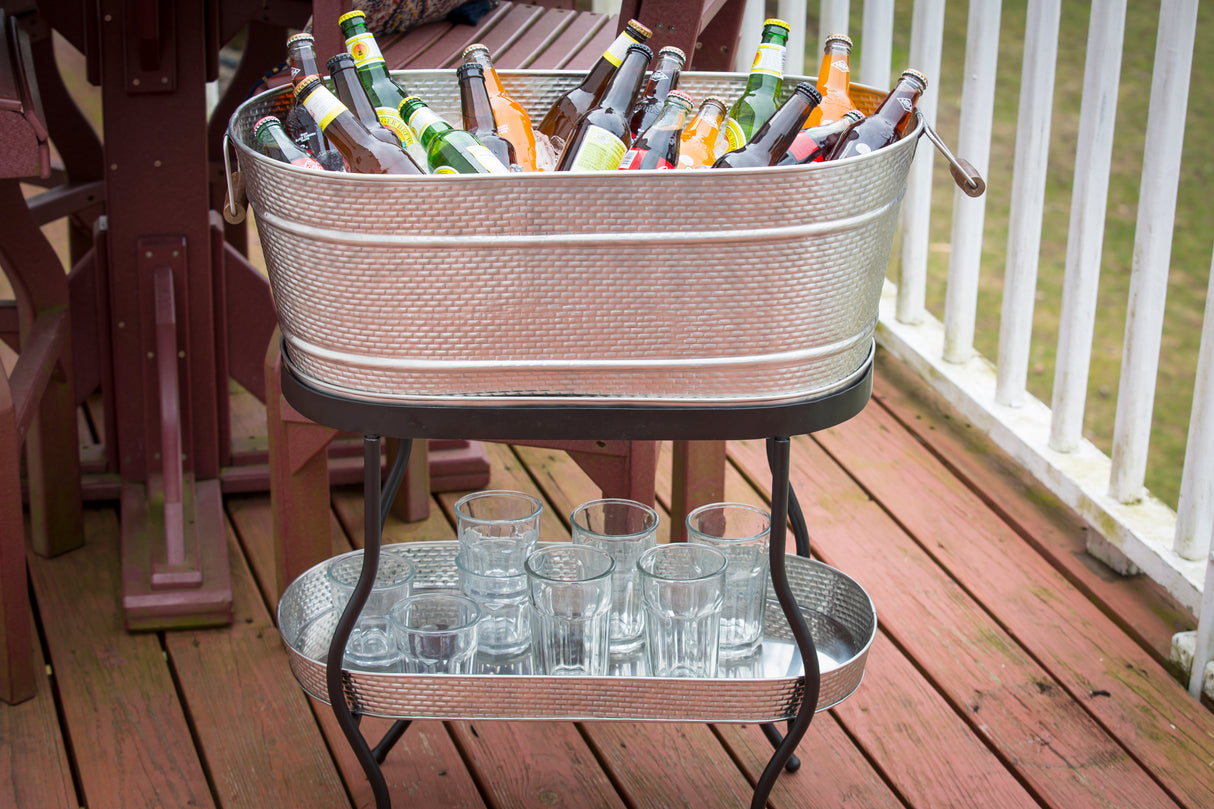 A Brickhouse Collection beverage tub set by TableCraft Products, filled with ice and assorted bottled drinks, sits atop a stand on the wooden deck; empty glass tumblers are neatly arranged on the lower tray shelf near the white railing.