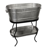 A large, oval-shaped, silver metal beverage tub with a hammered texture sits on a black TableCraft Products beverage tub stand. The stand features curved legs and supports a matching smaller tray below. The tub has a handle on one side.