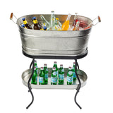 A metal beverage tub filled with ice and assorted bottled drinks sits on a black TableCraft Products Brickhouse Collection Replacement Stand. Below, a shelf holds green glass bottles of sparkling water. The tub features wooden handles against a plain white background.