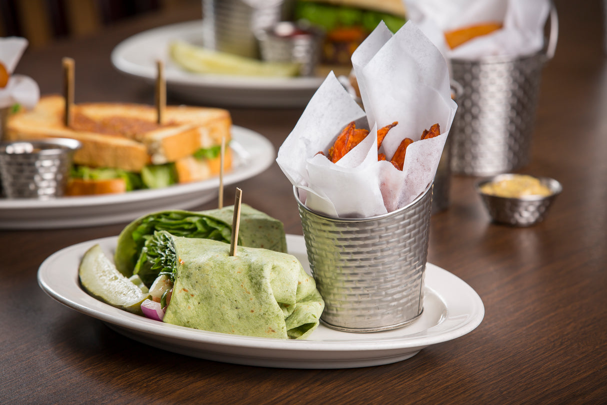 A spinach wrap filled with vegetables sits on a white plate, accompanied by a pickle spear. Sweet potato fries are served in a Brickhouse Collection Fry Cup, crafted from stainless steel, with a sandwich and more fries visible in the background.