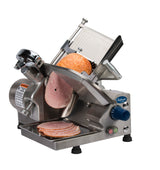 A stainless steel 12 inch slicer from the Chefmate Series with control knobs and safety labels slices a round ham into thin, even pieces that fall onto a tray below. This compact slicer is set against a plain white background.