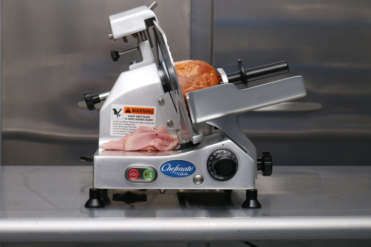 A Chefmate food slicer with a 12" knife sits on a metal counter, slicing thin pieces from a round loaf of ham. Stacked slices rest at its base, while the manual slicer features a warning label, power switch, and thickness dial.