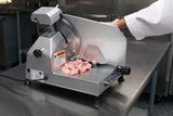 A person in a white coat uses a commercial 12" knife food slicer to cut pink deli meat in a stainless steel kitchen. The slicer has warning labels and controls, and thin slices of meat are stacked neatly on the machine’s tray.