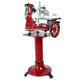 A vintage red and silver flywheel slicer stands on a matching red pedestal base. This Italian-designed slicer features a large hand-crank wheel, circular blade, and metal parts for securing and slicing meat, all set on a glossy platform.
