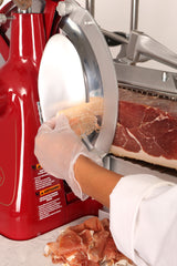 A person wearing a white glove expertly slices cured meat with a red and silver Italian-designed slicer. Thin slices fall onto a pile below, while the hand stays close to the sharp circular blade and visible safety labels.