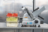 A Globe G12 12" manual slicer sits on a metal counter next to three stacked clear containers filled with sliced tomatoes, shredded lettuce, and sliced onions. The reflective metal surface in the background suggests an industrial kitchen setting.