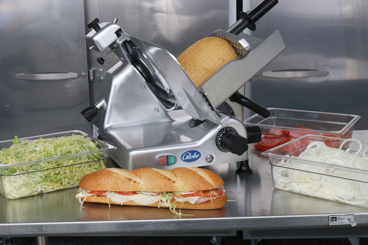 A Globe G12 12" manual food slicer holds a round loaf of cheese, surrounded by containers of shredded lettuce, tomato slices, and onion rings. Up front, a sub sandwich on a baguette overflows with deli meat and veggies against a stainless steel background.