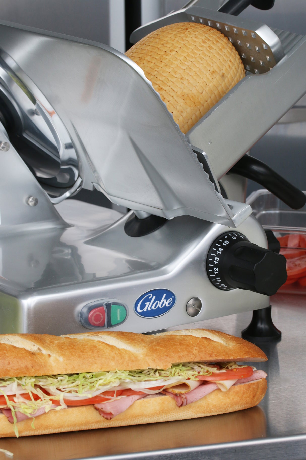 A Globe G12 12" manual slicer sits behind a fresh baguette sandwich filled with ham, lettuce, tomato, and cheese. The Globe food slicer displays its logo and control knobs, with sliced tomatoes in a clear container nearby.