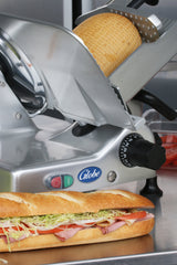 A Globe G12 12" manual slicer sits behind a fresh baguette sandwich filled with ham, lettuce, tomato, and cheese. The Globe food slicer displays its logo and control knobs, with sliced tomatoes in a clear container nearby.