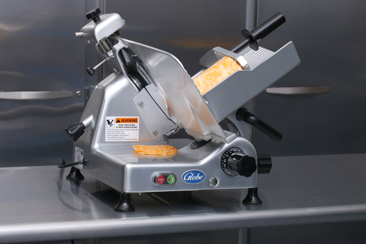 A Globe G12 12" manual food slicer with a dial, green and red buttons, and warning labels rests on a stainless steel counter. Slices and a block of marbled yellow and white cheese are on the tray, ready to be cut.