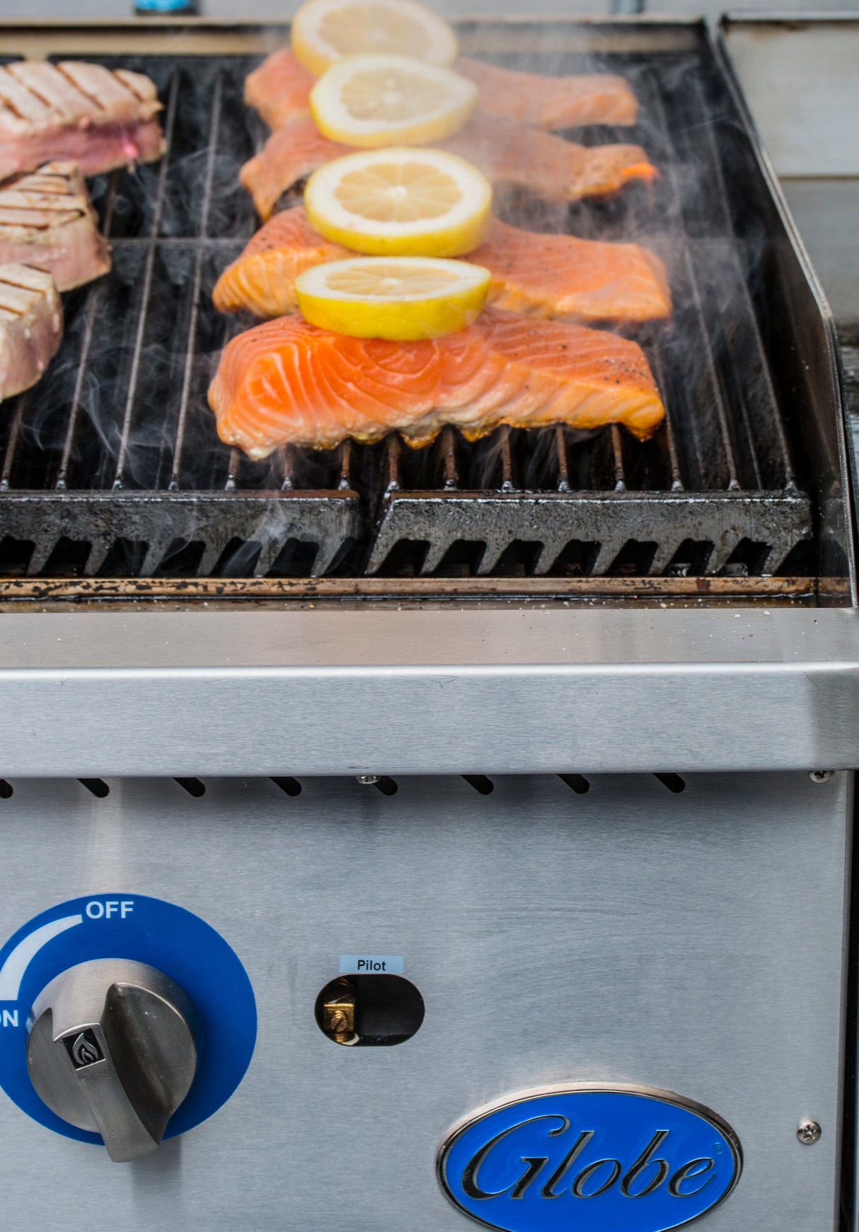 Three raw salmon fillets with lemon slices and two seared tuna steaks sizzle on a heavy duty charbroiler. The stainless steel appliance features a blue "Globe" logo and control knob up front, while steam rises from the grilling food.
