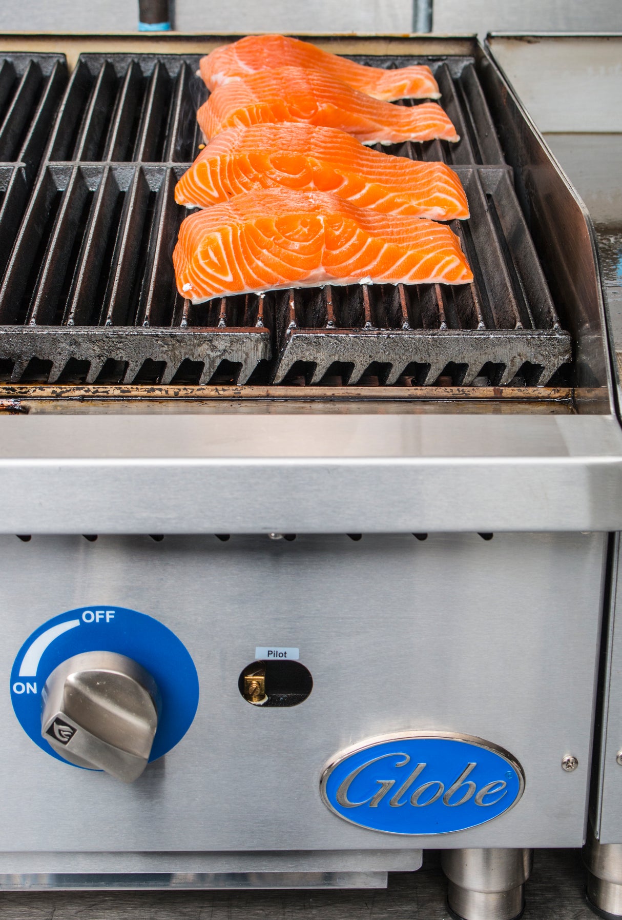 Four raw salmon fillets are lined up diagonally on a hot, black countertop charbroiler. Their bright orange color contrasts against the heavy duty cast-iron grates above a stainless steel control panel with a blue knob and the “Globe” logo.