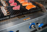 Close-up of a gas charbroiler with raw shrimp skewers, pink tuna steaks, and orange salmon fillets sizzling on heavy duty cast-iron grates. Blue temperature control knobs and a warning label are visible; steam rises from the fresh seafood.