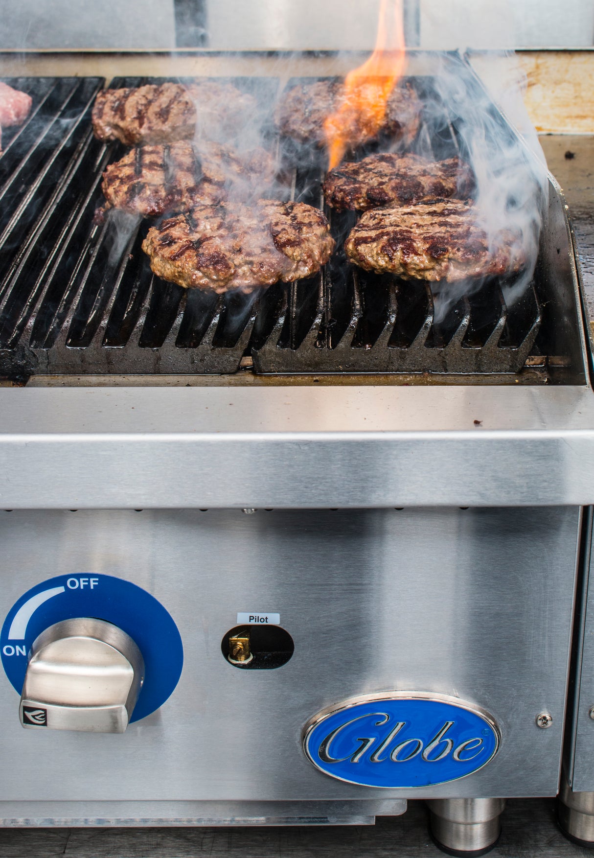 A gas charbroiler with heavy duty cast-iron grates cooks several beef burger patties, with flame and smoke rising. The stainless steel grill features a blue Globe logo, ignition switch set to Pilot, and a blue temperature dial marked OFF.