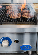 A gas charbroiler with heavy duty cast-iron grates cooks several beef burger patties, with flame and smoke rising. The stainless steel grill features a blue Globe logo, ignition switch set to Pilot, and a blue temperature dial marked OFF.