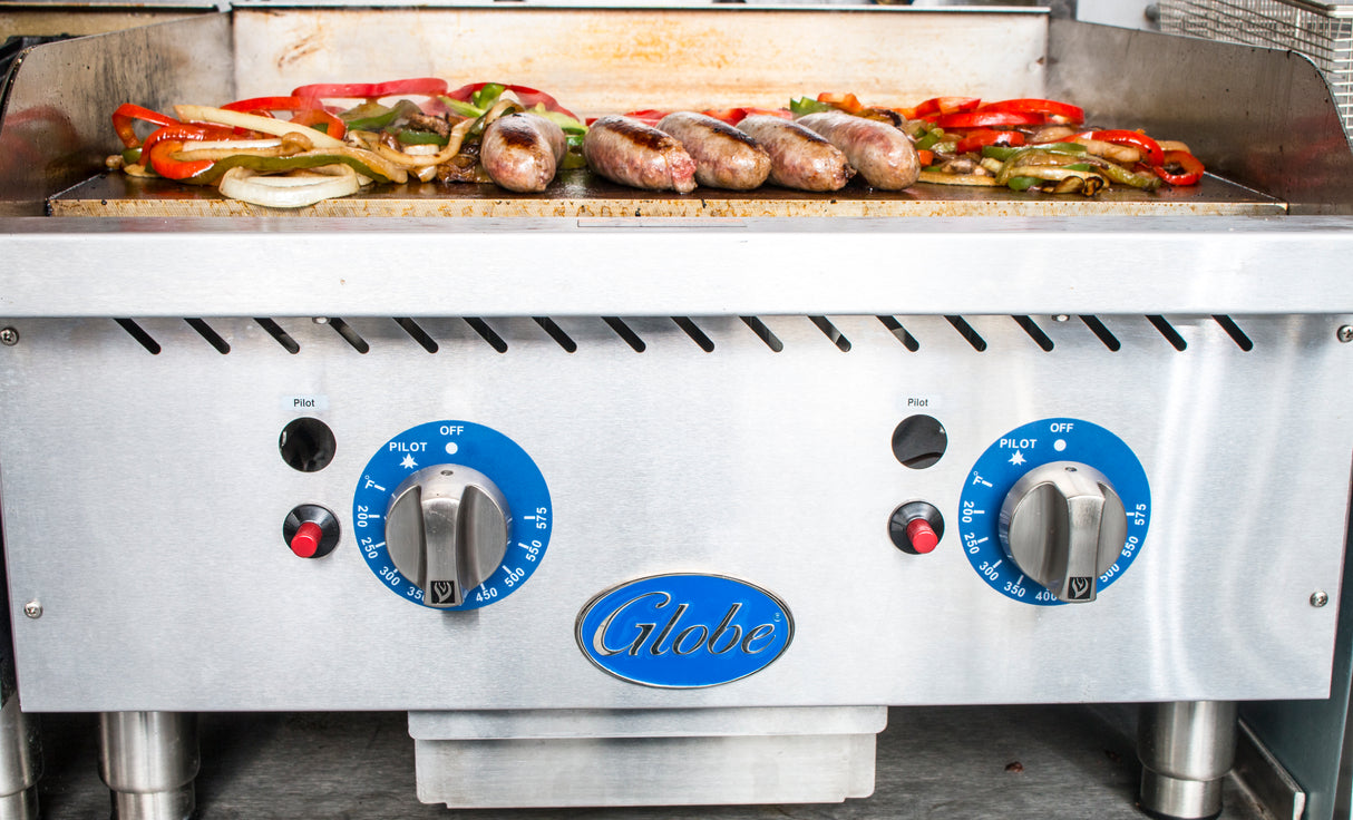 Five sausages and sliced onions, red, and green bell peppers are grilling on a stainless steel gas countertop griddle with blue Globe brand thermostatic controls set to medium. The vegetables and sausages are browning perfectly.