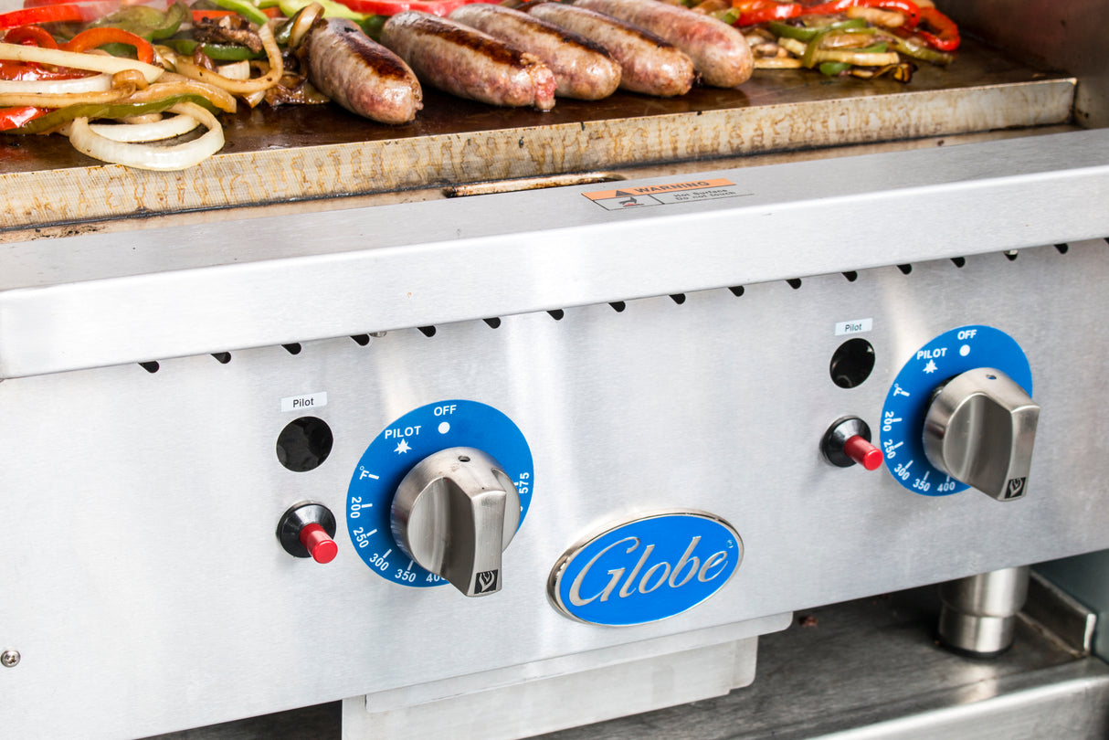 Sausages, onions, and bell peppers sizzle on a stainless steel Globe gas countertop griddle. Two blue thermostatic controls adjust the temperature settings as the food cooks perfectly on the hot surface.
