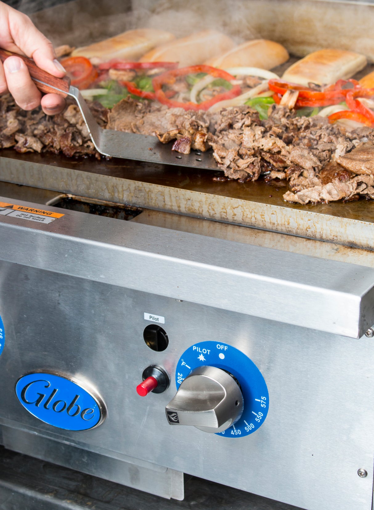 A close-up of a stainless steel gas countertop griddle cooking sliced beef, onions, and bell peppers, with sandwich rolls toasting in the background. A hand uses a spatula to stir the meat near Globe’s large blue thermostatic controls.