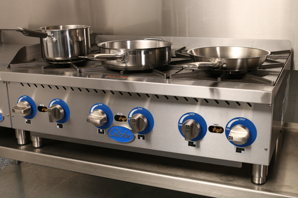 A commercial stainless steel Globe GHP36G stove features four burners with blue and silver knobs. Three pots and pans sit atop this gas countertop hotplate, branded “Globe,” in a spotless kitchen setting.