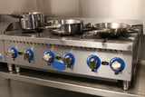 A commercial stainless steel Globe GHP36G stove features four burners with blue and silver knobs. Three pots and pans sit atop this gas countertop hotplate, branded “Globe,” in a spotless kitchen setting.