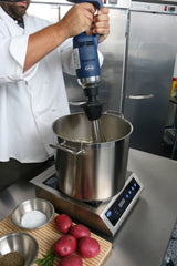 A chef in a white coat uses an Immersion Blender with a removable stainless steel shaft to blend food in a metal pot on an induction cooktop. On the counter are red potatoes, rosemary, salt, and pepper. Stainless steel refrigerators are in the background.