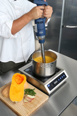 A chef in a white coat uses a variable speed Immersion Blender with a removable stainless steel shaft to mix soup in a pot on an induction cooktop. A halved butternut squash, sliced vegetables, and herbs sit on a wooden cutting board nearby.