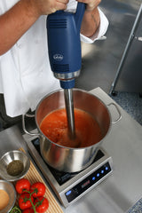 A chef uses a variable speed Immersion Blender with a 14" stainless steel shaft to puree red tomato soup in a stainless steel pot on an induction cooktop. Fresh tomatoes, herbs, and spices are arranged nearby; only the chef's arms and hands are visible.