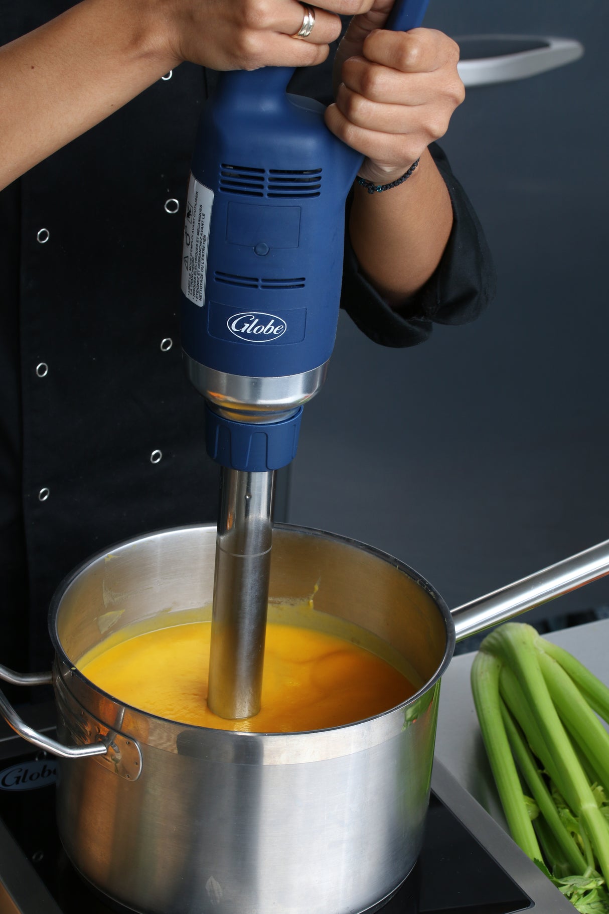 A person uses a large blue Immersion Blender with a 14" stainless steel shaft to blend bright yellow soup in a pot on the stovetop. The person's hands are visible, and celery stalks rest nearby. The background is out of focus.
