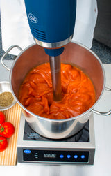 A large Immersion Blender with a 14" stainless steel shaft and variable speed mixes bright red tomato sauce in a pot on an electric stove. Fresh tomatoes, a metal tray with dried herbs, and a wooden board sit nearby. A person in white stands behind.