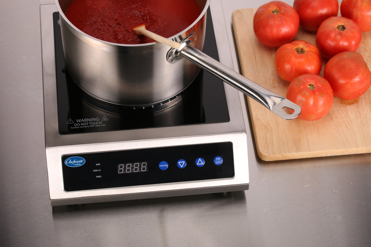 A pot of tomato sauce simmers on a countertop electric Induction Range with a wooden spoon inside. Nearby, six whole tomatoes rest on a cutting board atop the stainless steel construction countertop, with the cooktop’s digital display and buttons visible.