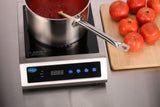 A pot of tomato sauce simmers on a countertop electric Induction Range with a wooden spoon inside. Nearby, six whole tomatoes rest on a cutting board atop the stainless steel construction countertop, with the cooktop’s digital display and buttons visible.