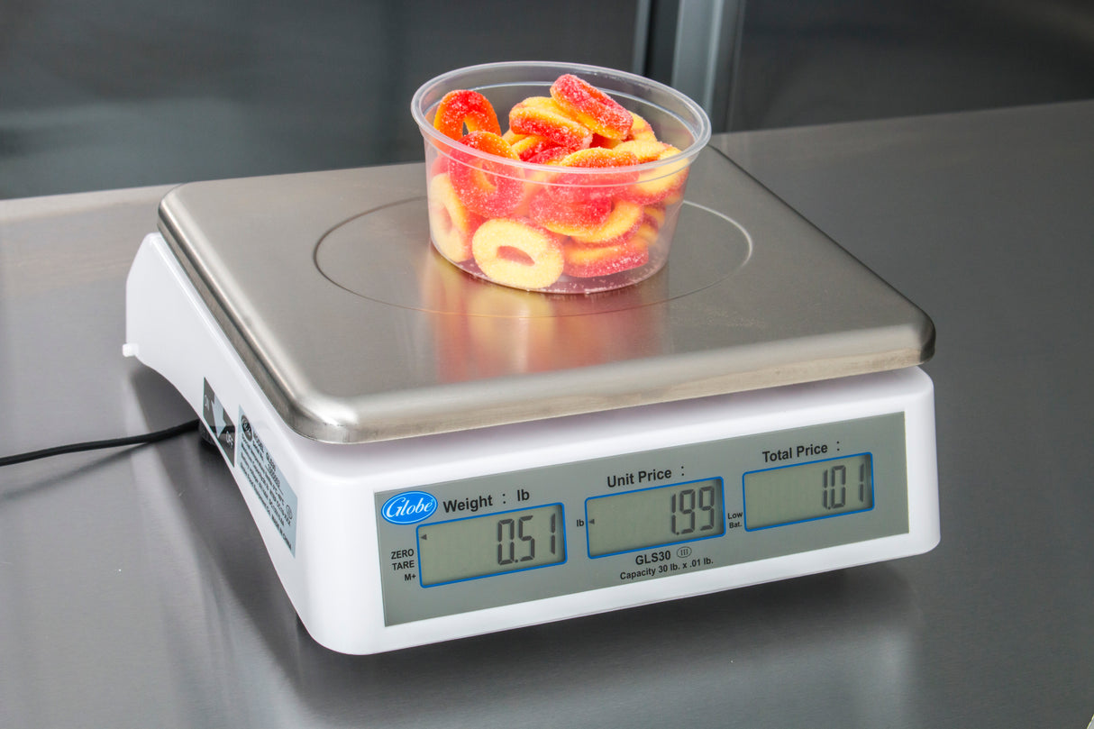 A clear plastic container filled with red and yellow candy rings sits on a price computing scale with a dual LCD display. The scale, legal for trade, shows a weight of 0.51 lb, unit price of 1.99, and total price of 1.01 on the stainless steel countertop.