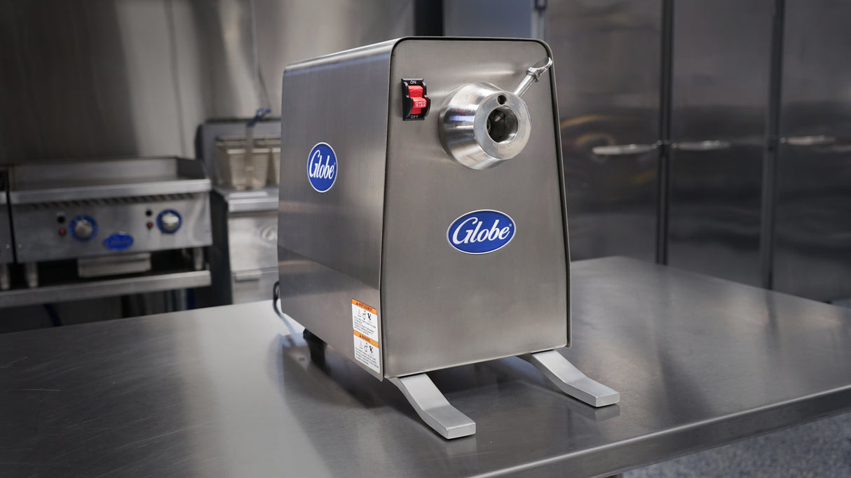 A stainless steel Globe brand meat grinder with 1 HP Power Drive Unit is on a reflective metal countertop in a commercial kitchen, with appliances and cabinets in the background. The grinder has a round feed tube, on/off switch, and stands on two sturdy legs.
