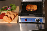 A grilled sandwich is toasted in a panini grill. On a wooden board nearby are slices of bread, leafy lettuce, tomato, onion, and pepperoni atop a stainless steel countertop. The panini grill's control panel and cast iron grooved plates are visible in front.