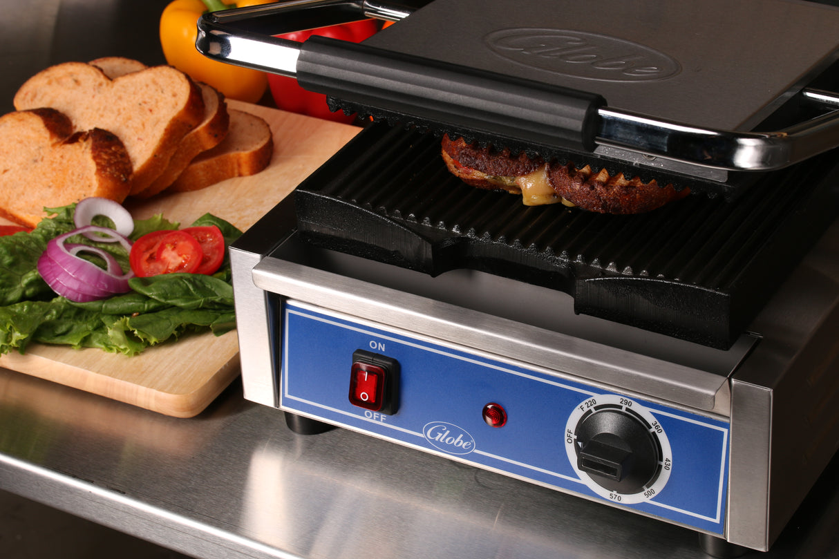 A sandwich is grilling in a panini grill with melted cheese visible. Nearby, a wooden board holds sliced bread, lettuce, tomato, onion, and a yellow bell pepper. The scene is set on a stainless steel countertop.