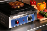 A close-up of a countertop electric grill with cast iron grooved plates grilling a sandwich with melted cheese. The blue control panel features a red on/off switch and temperature dial. In the background are fresh vegetables, bread, and sandwich ingredients.