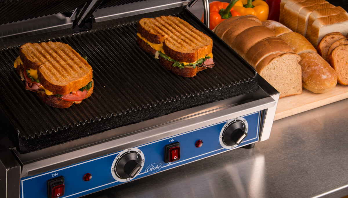 A close-up of two panini sandwiches grilling on a countertop electric grill. Behind the panini grill are sliced and whole loaves of bread, with bell peppers resting on a wooden cutting board atop the metallic countertop. The grill is turned on and heating.