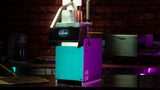 A Globe cheese shredder with a stainless steel upper section and teal lower body stands on a dark reflective countertop. Designed for high volume use, it features continuous feed capability, while purple and teal highlights illuminate the blurred kitchen backdrop.