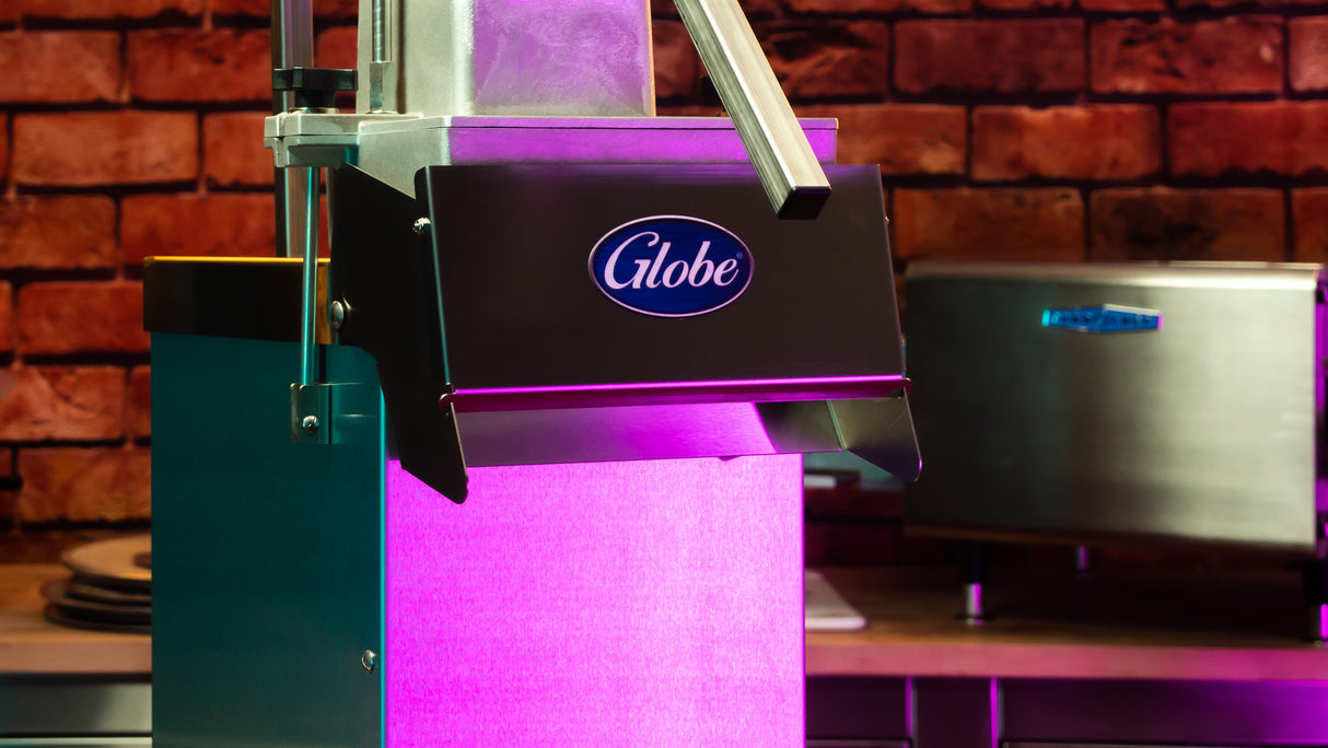 Close-up of a Globe brand commercial kitchen slicer with a metallic handle, illuminated by bright pink and purple light. Ideal for high volume operations, it stands on a countertop before a brick wall, near another appliance.