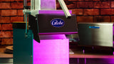 Close-up of a Globe brand commercial kitchen slicer with a metallic handle, illuminated by bright pink and purple light. Ideal for high volume operations, it stands on a countertop before a brick wall, near another appliance.