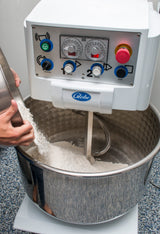A person pours flour into a large spiral dough mixer with a 130 lbs capacity. The industrial mixer features a stainless steel bowl, visible mixing arm, control panel with dials and buttons, red emergency stop, and the “Globe” brand on the front.