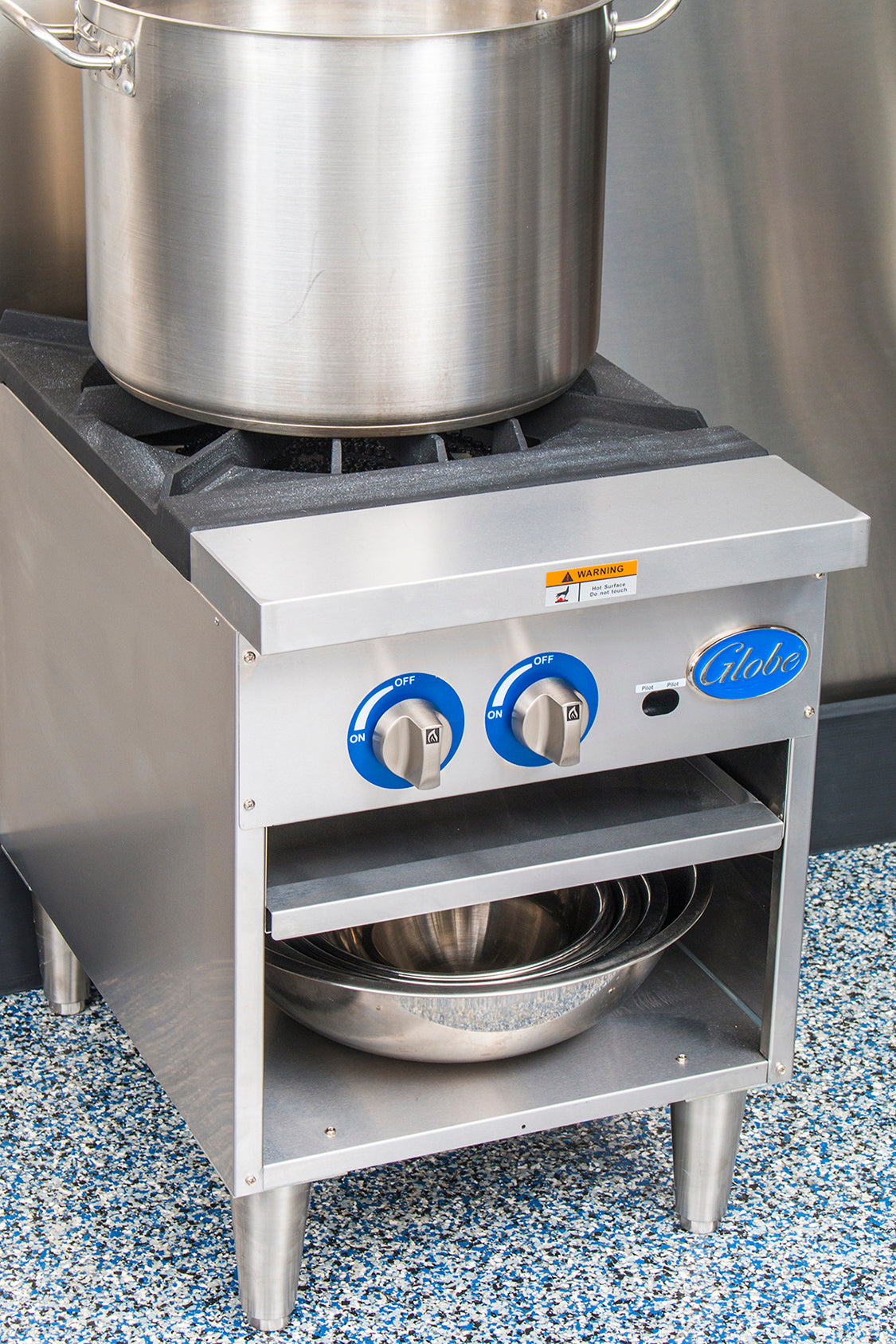 A large stainless steel pot sits on a Globe GSP18G gas stock pot range with two blue control knobs. Below, two nested metal mixing bowls are stored on a shelf. The stove stands on four metal legs on a blue speckled floor.