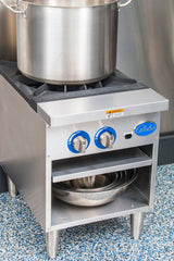 A large stainless steel pot sits on a Globe GSP18G gas stock pot range with two blue control knobs. Below, two nested metal mixing bowls are stored on a shelf. The stove stands on four metal legs on a blue speckled floor.