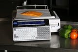 A label printing scale on a stainless steel counter displays sliced cheese. Its dual-sided LCD display shows weight and price details. Beside the scale are a green bell pepper, green chili pepper, and red tomato, as a receipt prints out.